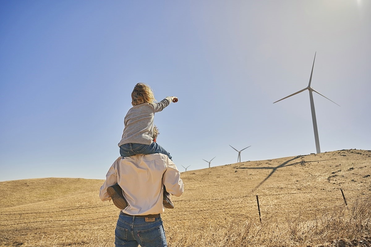 A child on a parent's shoulders pointing towards wind turbines in a dry, open landscape.