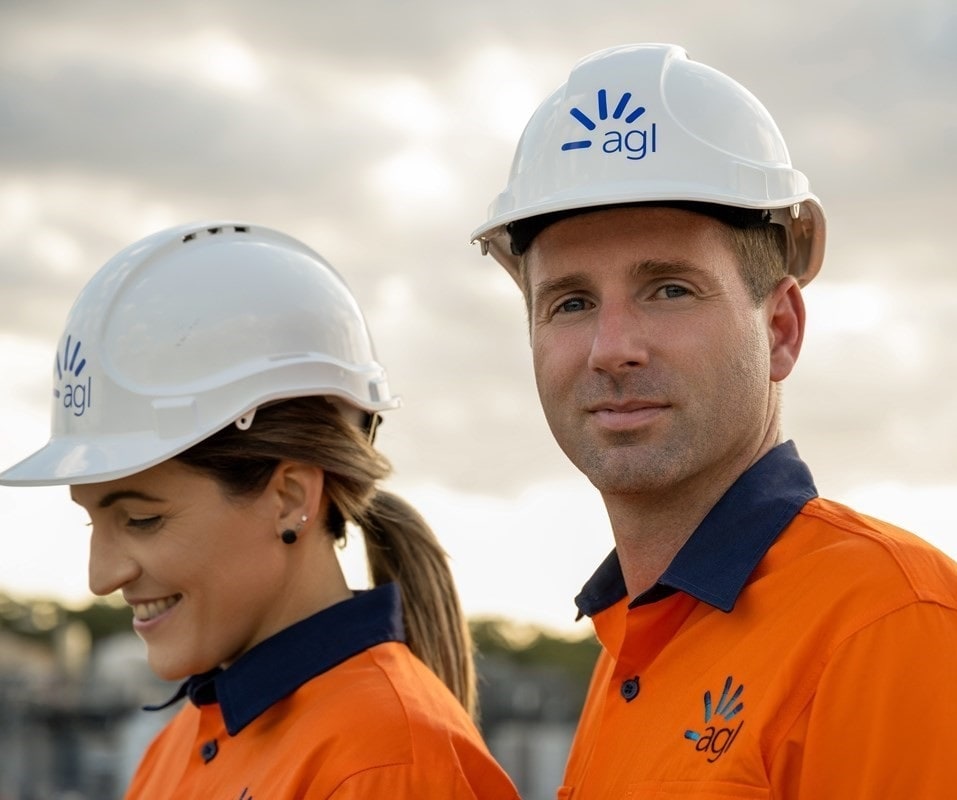 Two workers in AGL hard hats, one man in focus, one woman smiling ahead.