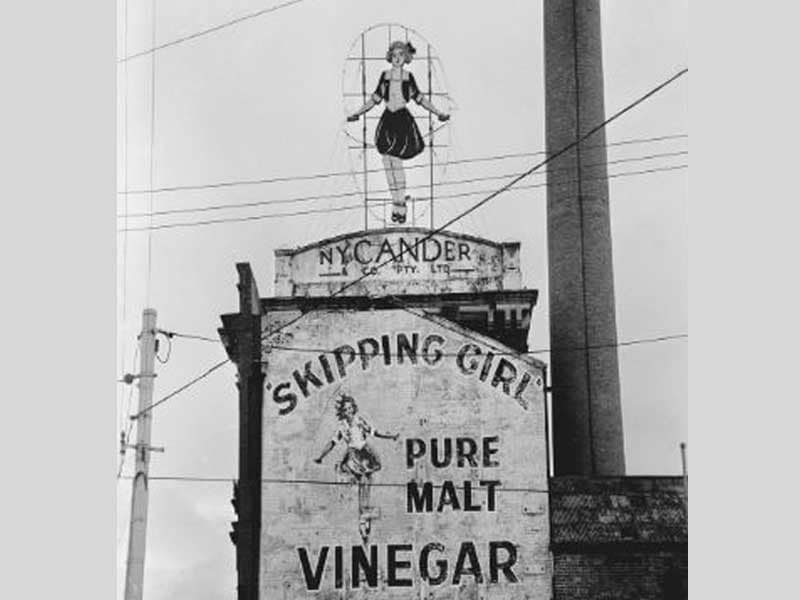 Historic photo of 'Skipping Girl' vinegar sign with neon outline in black and white.