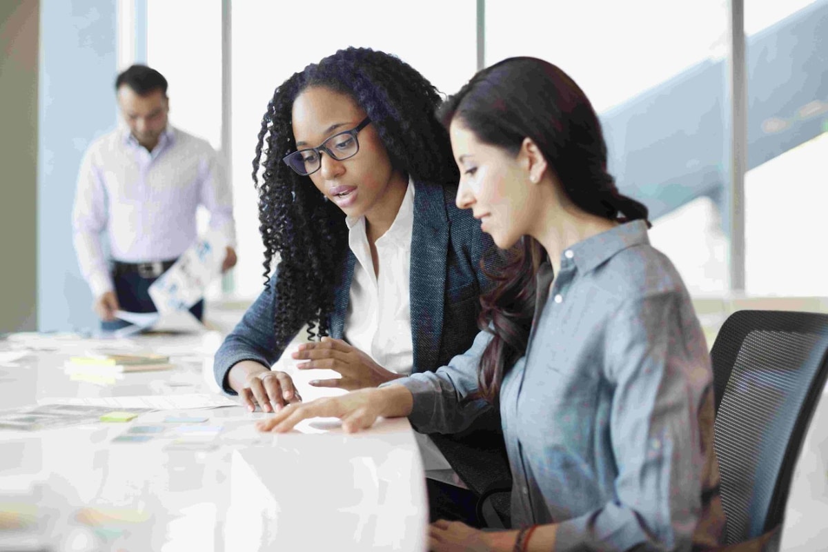 Two corporate women confer at a desk with a man flipping through papers in the background