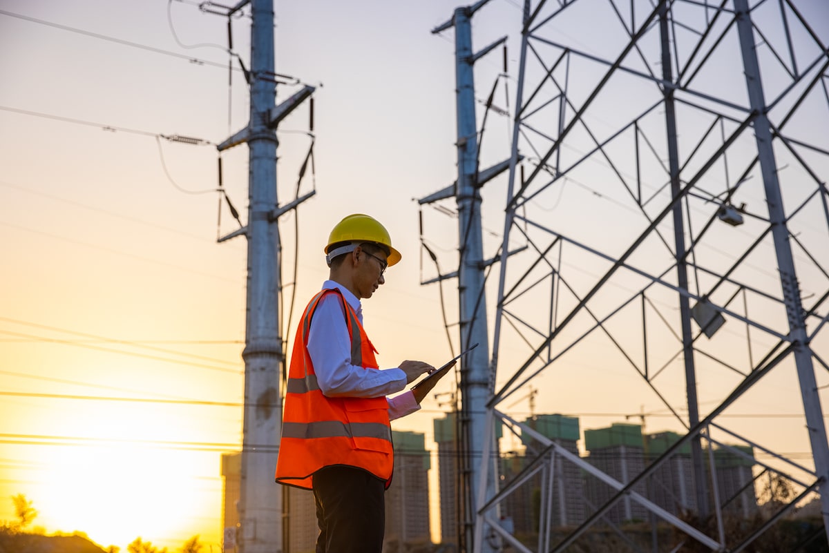 Tradie wearing an orange high vis vest and yellow safety helmet standing next to a pylon at a sub-station