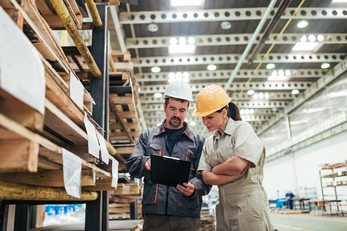 Two people standing next to large shelves in a warehouse, looking at a clipboard