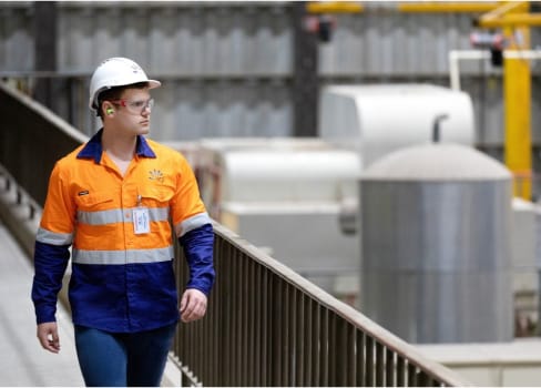 A man wearing a high vis vest and a hard hat walking along a catwalk overlooking a production plant