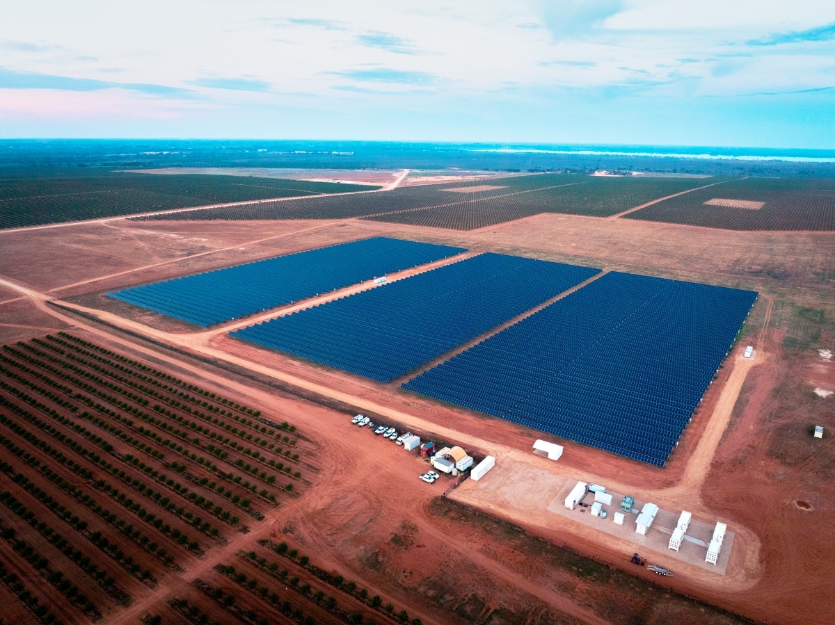 Solar farm with rectangular panels in rural fields; vehicles and buildings visible.