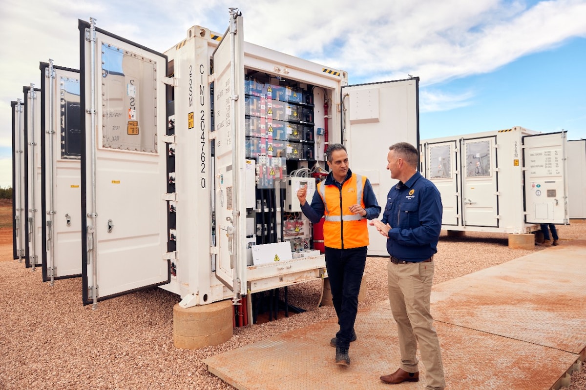 Two men stand in front of an open shipping container with electrical equipment at an outdoor site.