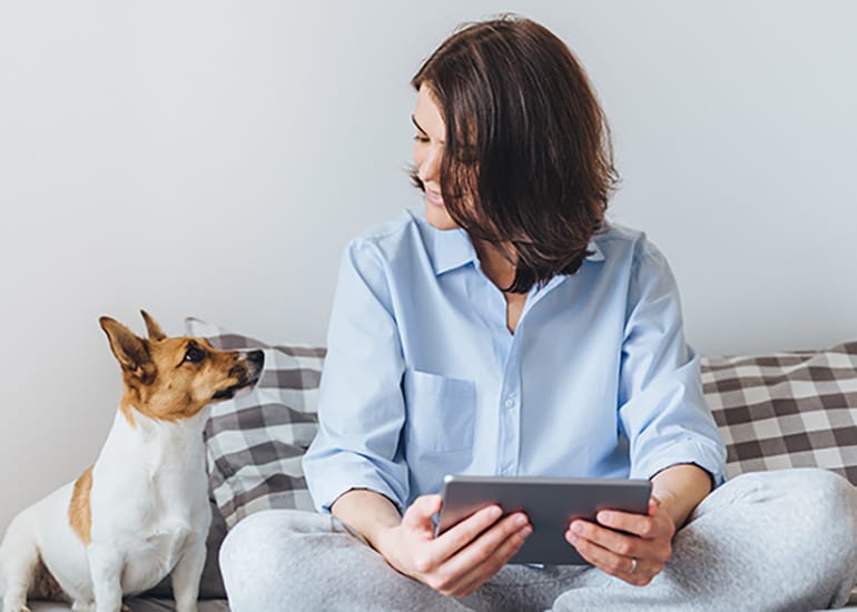 Woman with a tablet device sits on the bed with her dog.