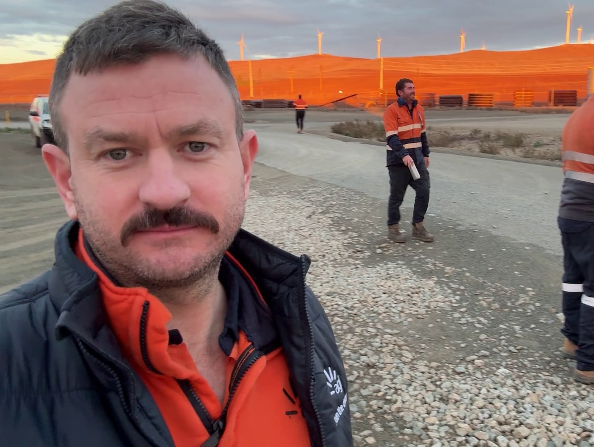 Photo of Matt in high vis and jacket standing in front of wind farm