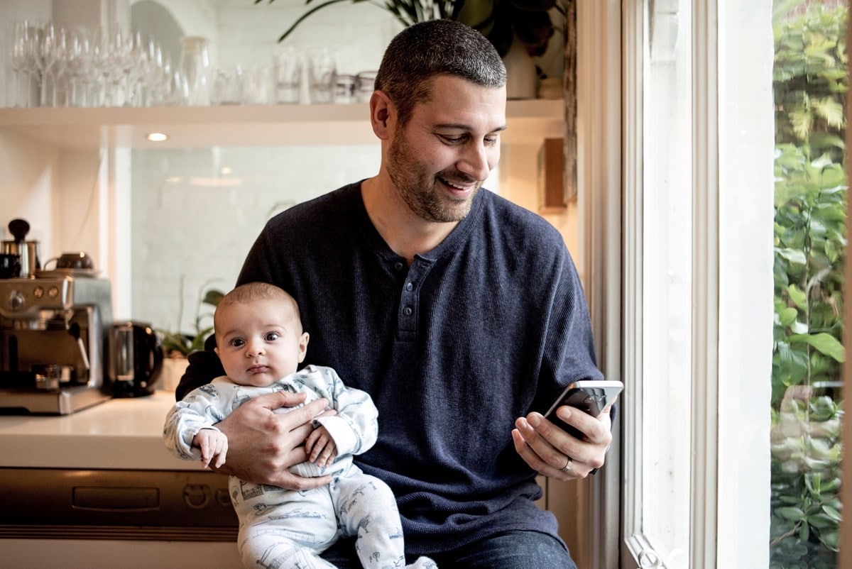 Man in kitchen holding a baby while using a mobile device