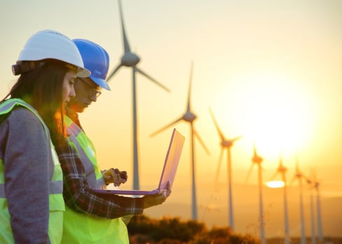 Two coworkers in high vis working on laptop in a wind turbine farm while the sun sets 