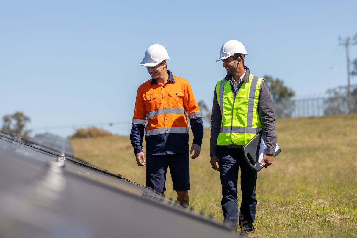 Close up of two men in high vis vests and hard hats walking through a solar farm inspecting solar panels