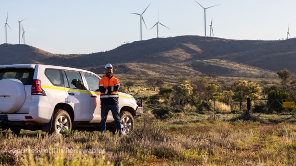 A man in high vis and a hard hat standing beside a white SUV with a wind turbine farm on a hill in the background