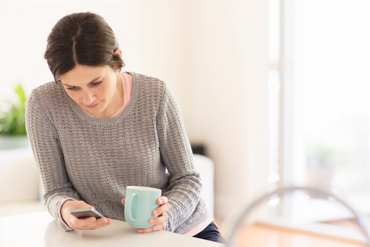A young aged woman is reading from her mobile phone while drinking her morning coffee at home.