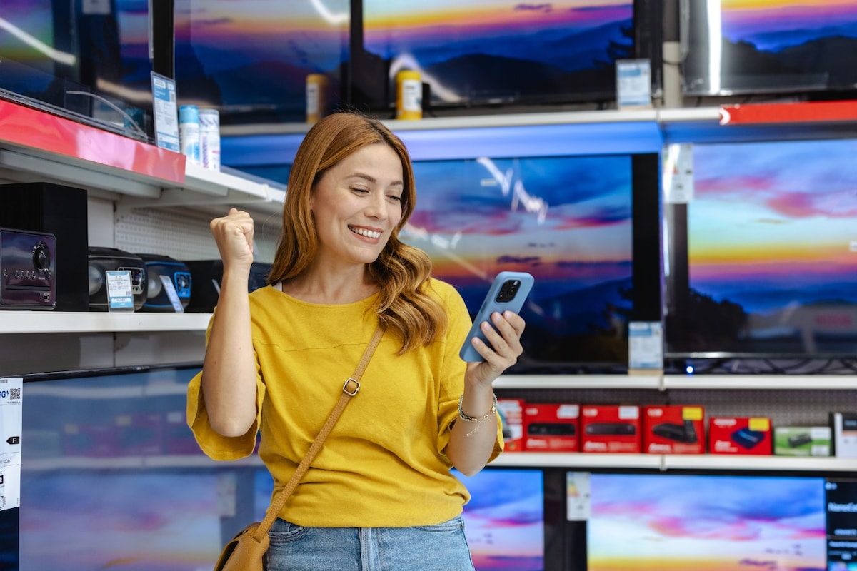A joyful woman holding a smartphone, celebrating a successful online purchase while standing in an electronics store, surrounded by TVs.