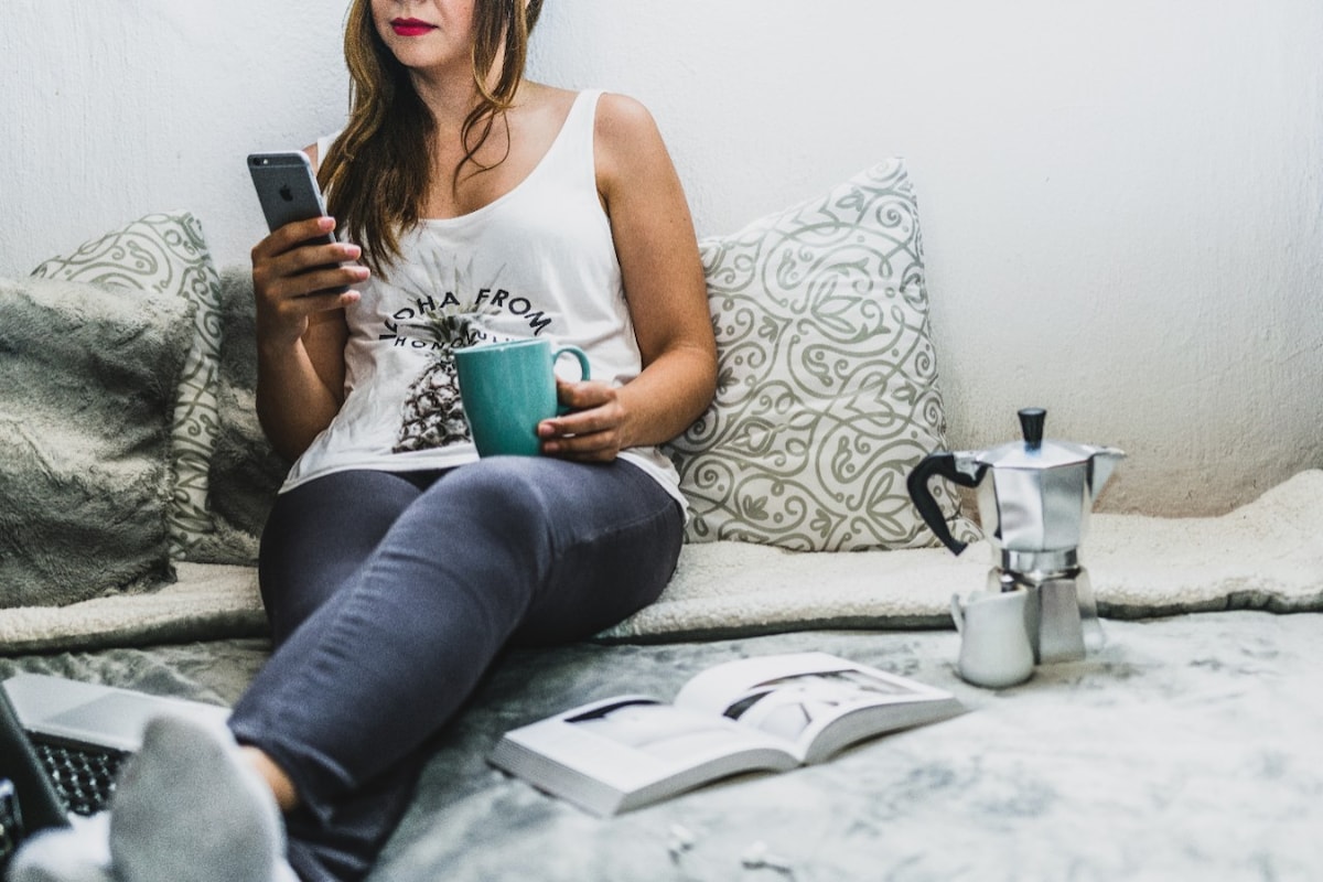 Woman sitting on bed with phone drinking coffee