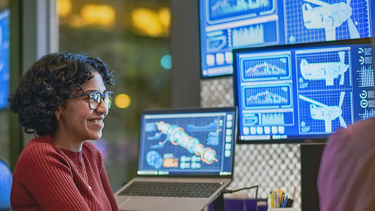 An individual in a red shirt is seated in front of multiple monitors displaying graphs and data in a design space..