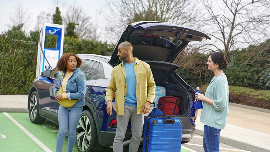 A family standing in front of an EV. The boot is open and camping gear packed inside.