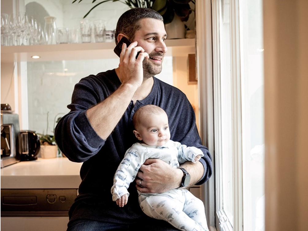 Man in kitchen holding a baby while using a mobile device